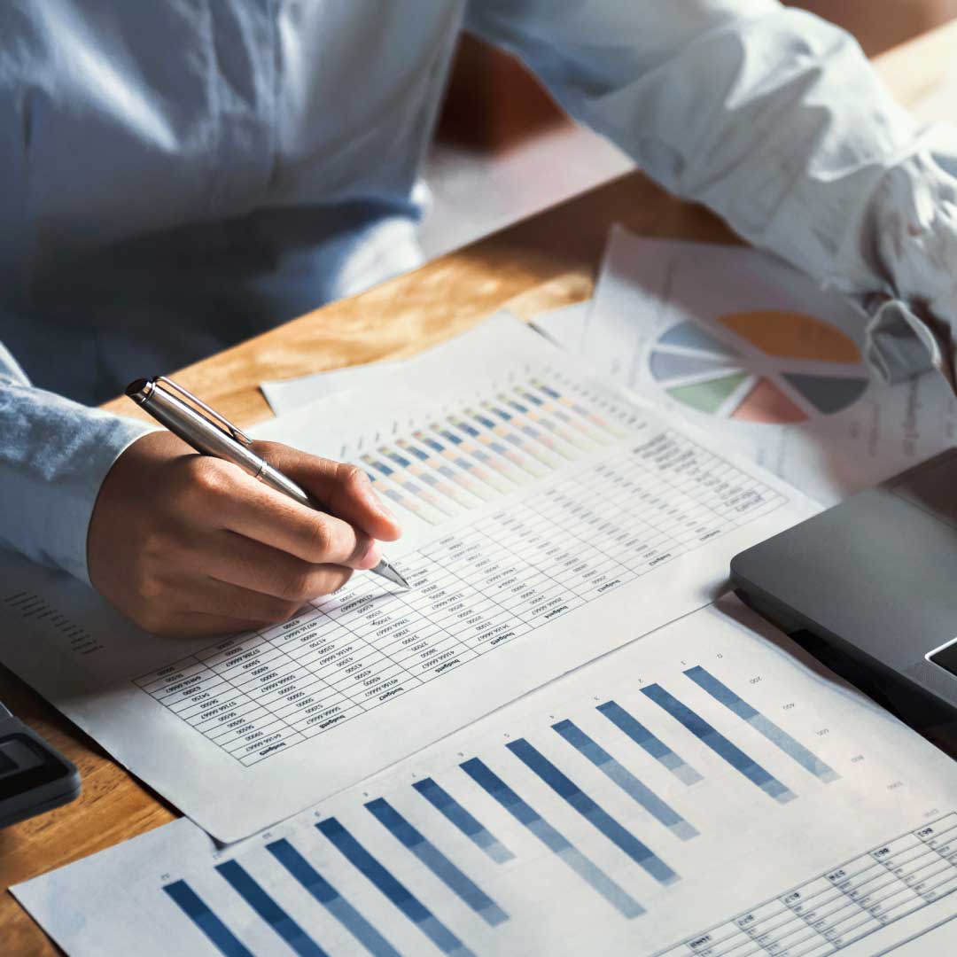 A man's hand holds a pen, pointing at a printed page showing a spreadsheet, sitting on a desk with other documents displaying financial data.