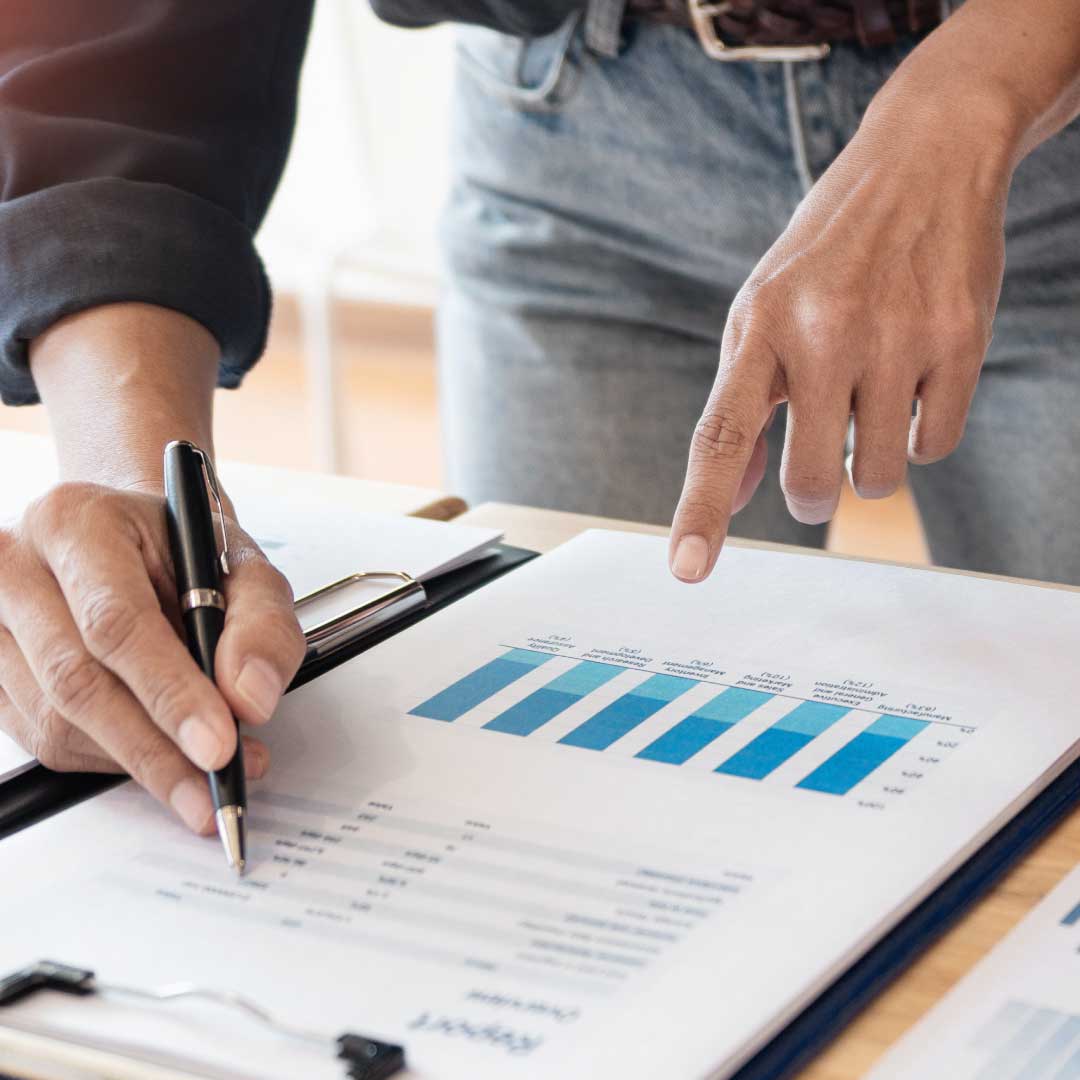 A man wearing blue jeans holds a pen, pointing at a page of financial data held on a clipboard.