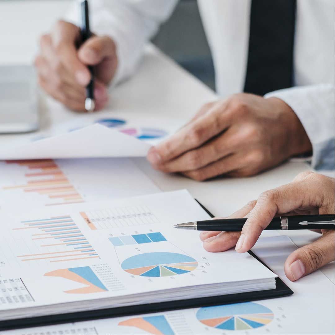 Two men are sitting at a table with pens in their hands, pointing at printed pages of financial data.