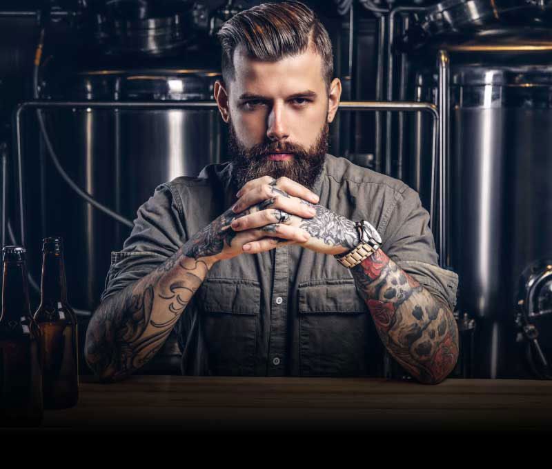 A bearded man with tattoos on his arms, sitting with his hands crossed and elbows on a bar, with beer bottles nearby and large silver stills in the background.