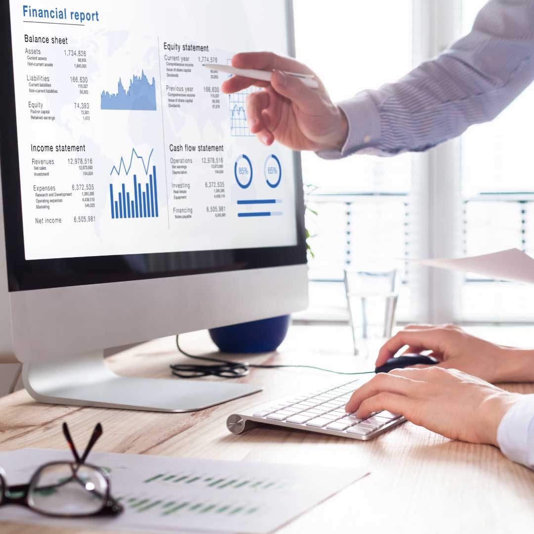 One man's hand points to financial data on a computer monitor, while another man's hands rest on the keyboard, ready to type.