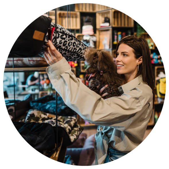 A woman holds her pet dog as she browses a pet store and checks out a tag on a leopard skin patterned pillow.